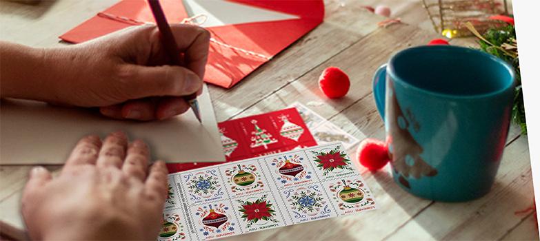 A person addressing a letter with First-Class Mail Forever® holiday stamps on the table.